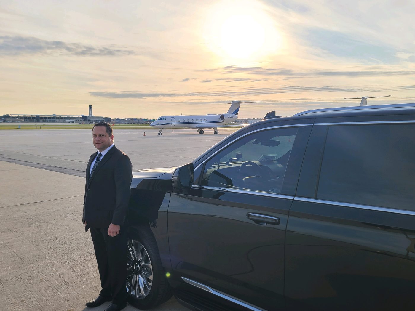 Chauffeur in a dark suit standing beside a black Cadillac Escalade ESV on the ramp at Milwaukee Mitchell, a private jet parked behind them at sunrise