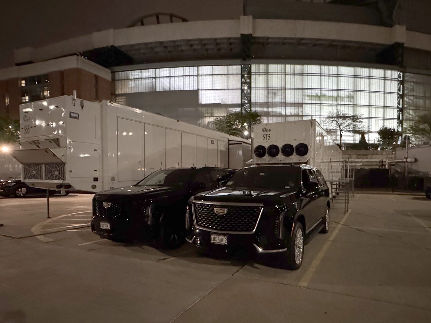 Two black Cadillac Escalade ESVs parked beside broadcast trucks outside the lit glass wall of American Family Field at night