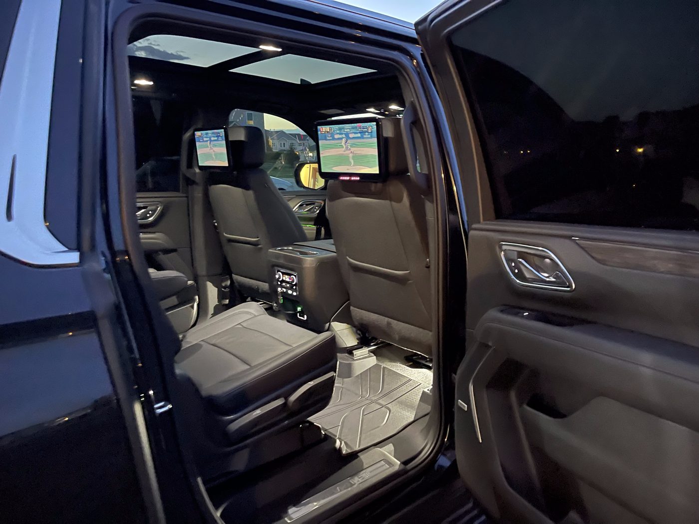 Second row of a Cadillac Escalade ESV at dusk with the rear door open, both rear screens showing a baseball game and the panoramic sunroof open