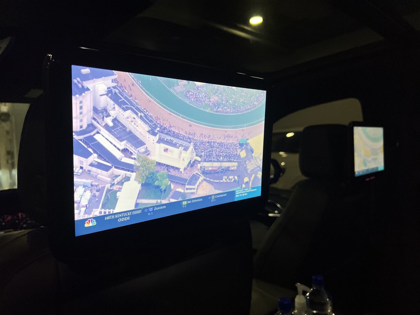 A rear-seat display in a Cadillac Escalade ESV showing a live broadcast, with a second screen visible beyond it