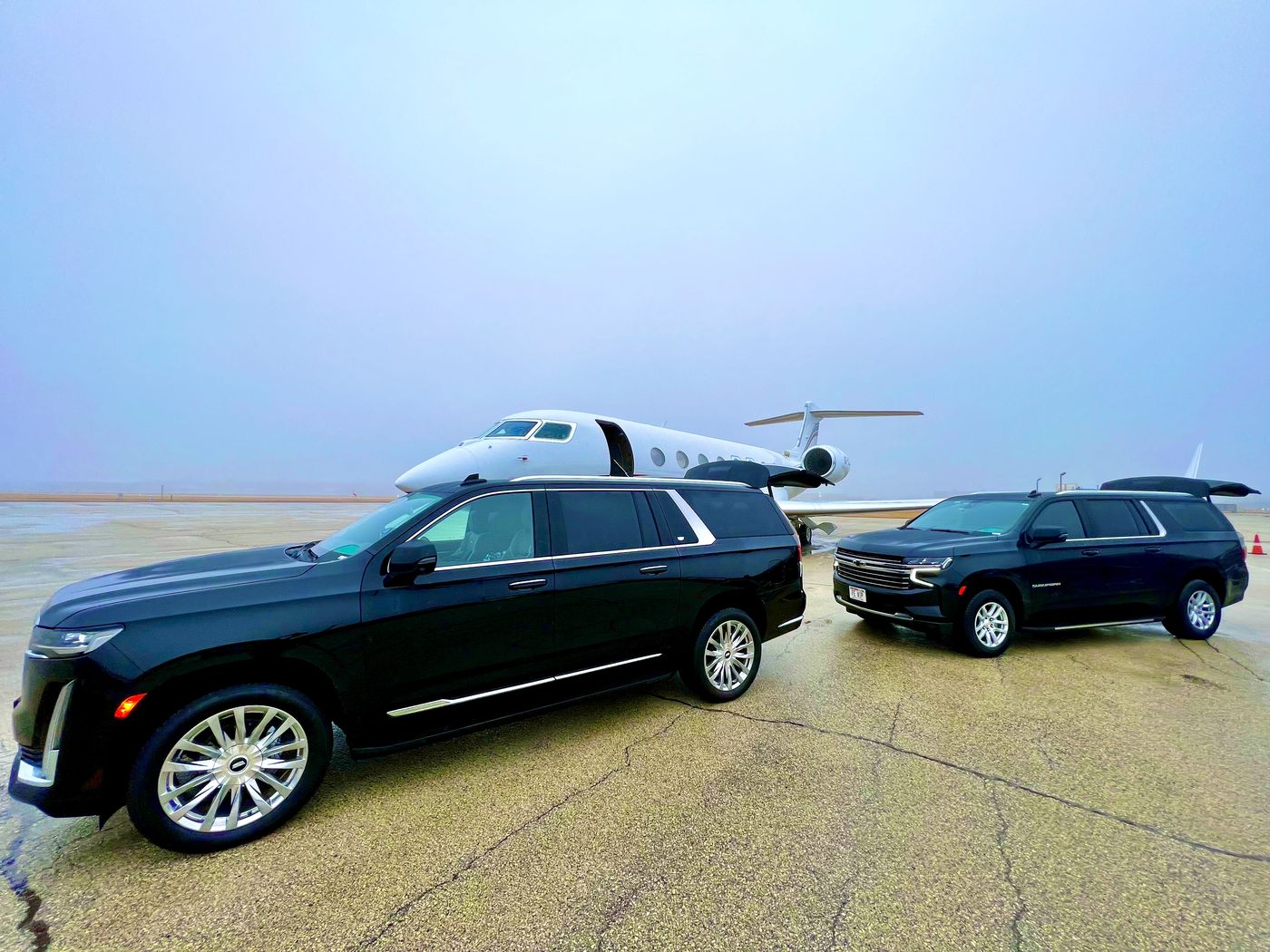 Black Cadillac Escalade ESV beside a private jet on the FBO ramp, meeting an arrival bound for Elkhart Lake