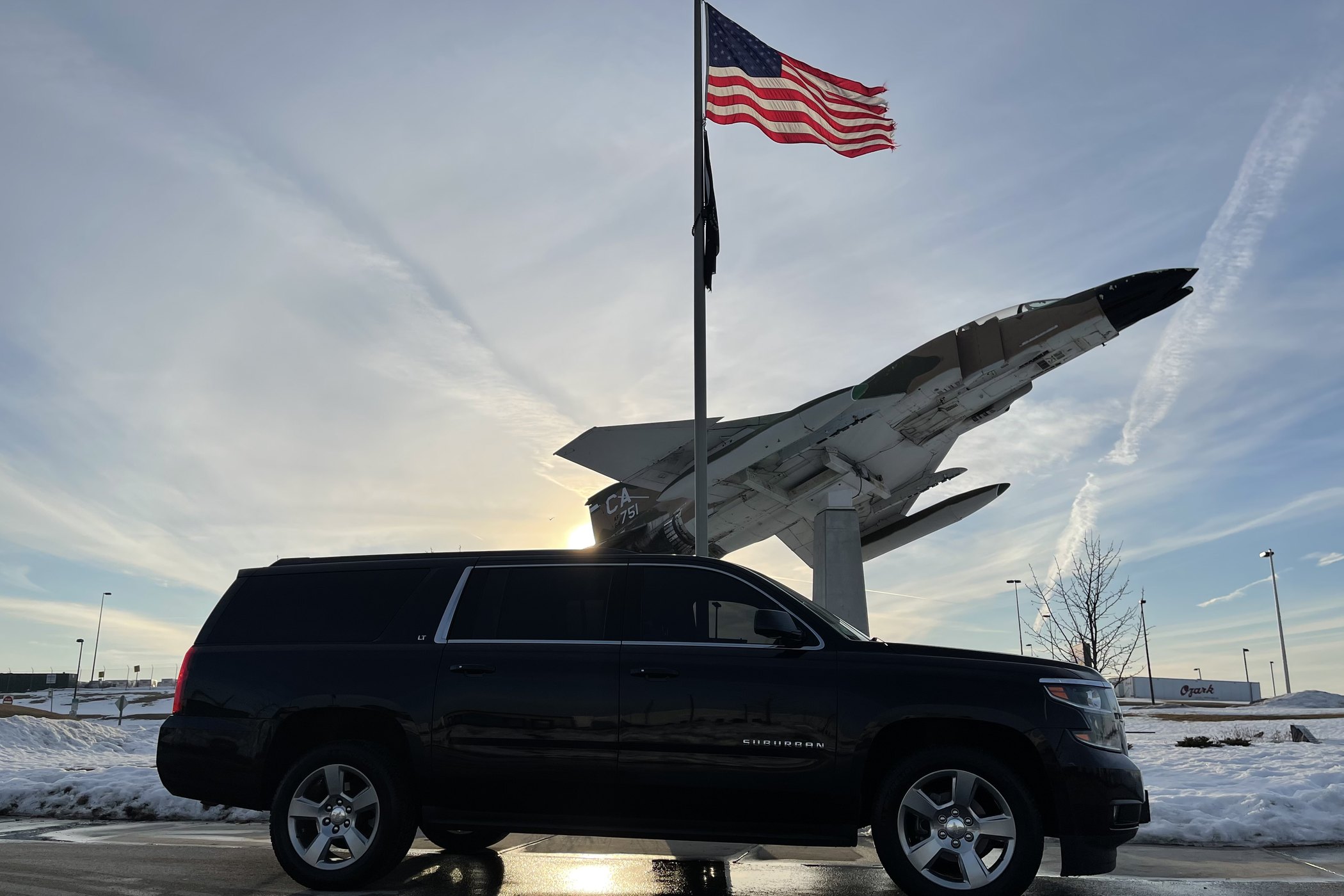 Black Chevrolet Suburban parked below the American flag and a display aircraft at Milwaukee Mitchell International Airport on a winter morning
