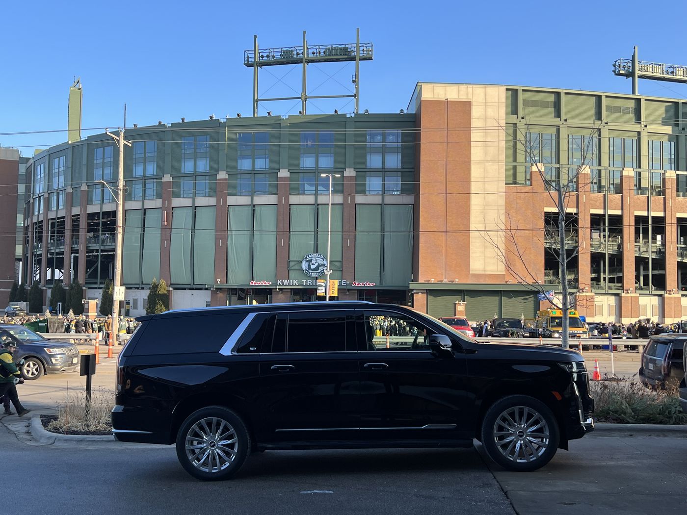 Black Cadillac Escalade ESV at the curb across from Lambeau Field on a cold afternoon, fans in green walking toward the gates
