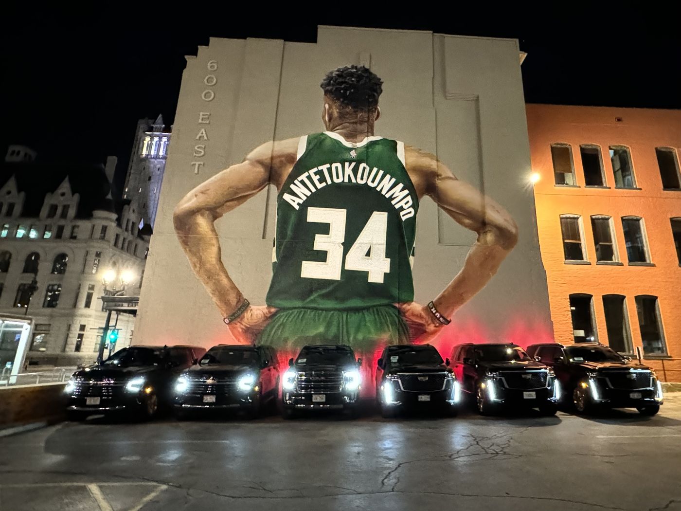 A line of black SUVs with headlights on, staged in front of a large mural on a building in downtown Milwaukee