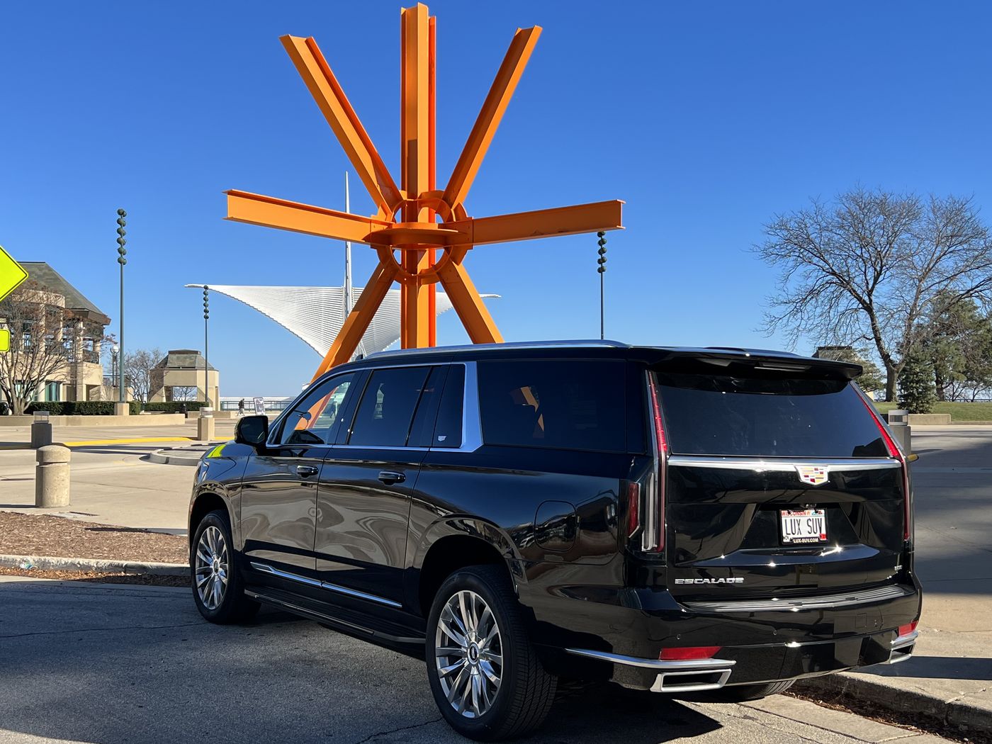 Black Cadillac Escalade ESV parked on the Milwaukee lakefront, with the orange steel of Mark di Suvero's Calling sculpture and the white brise soleil of the Milwaukee Art Museum behind it