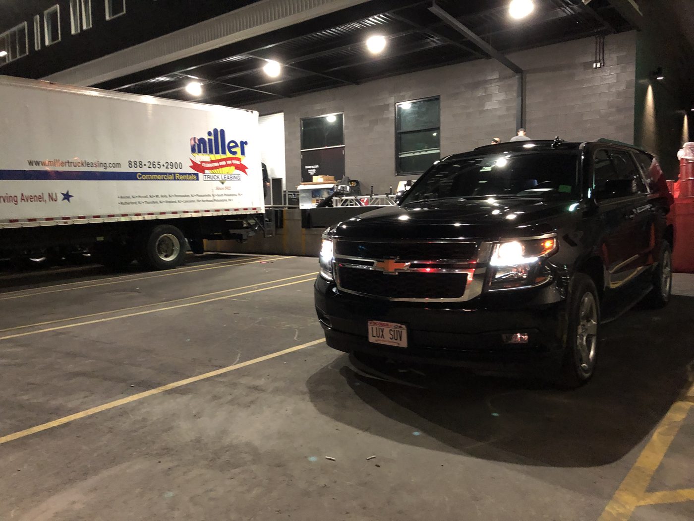 Black Chevrolet Suburban waiting at a backstage loading dock at night, a production truck being unloaded alongside it