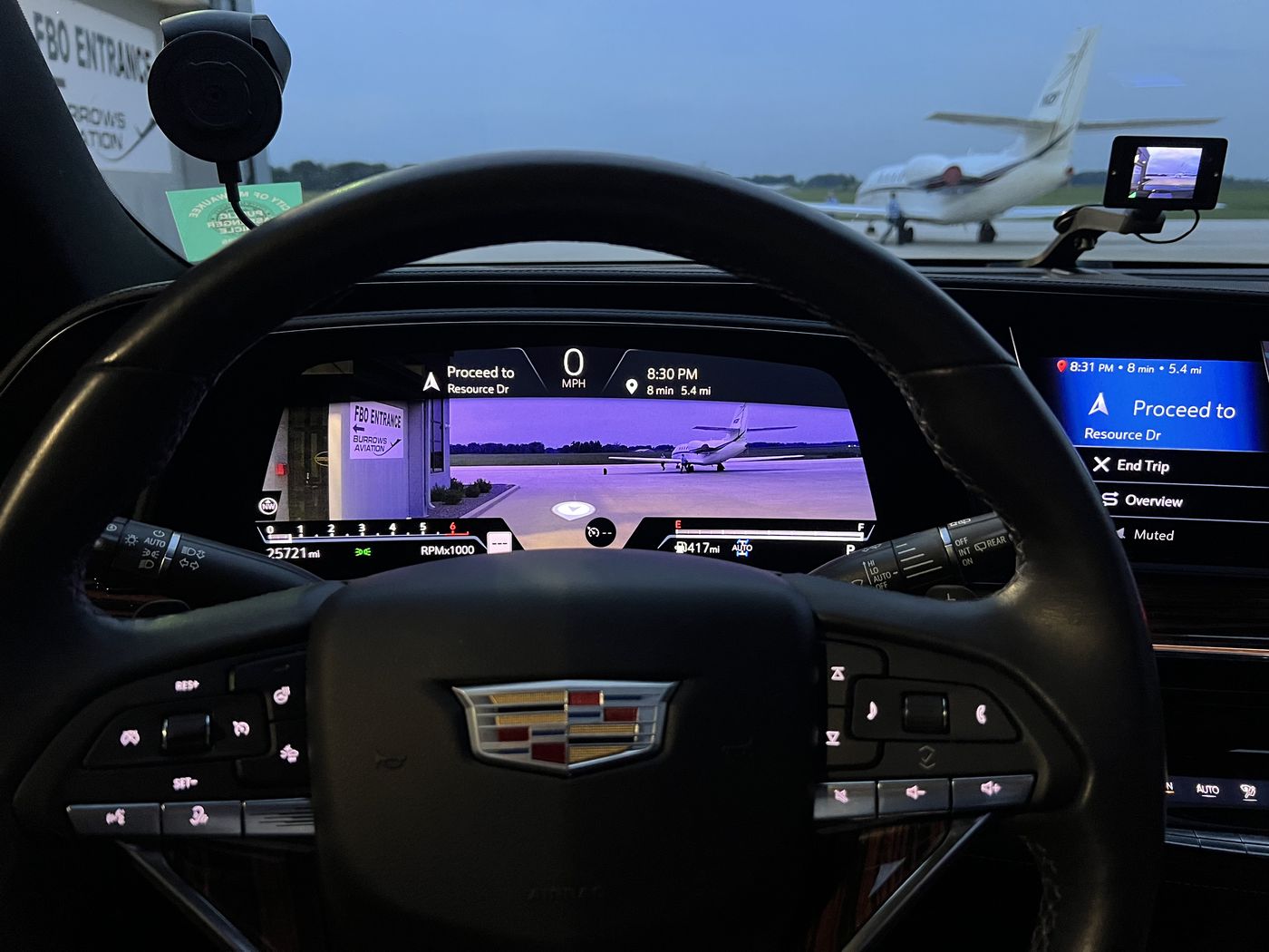 View from the chauffeur's seat of a Cadillac Escalade parked at an FBO entrance at dusk, a private jet on the ramp ahead and the navigation showing the trip complete