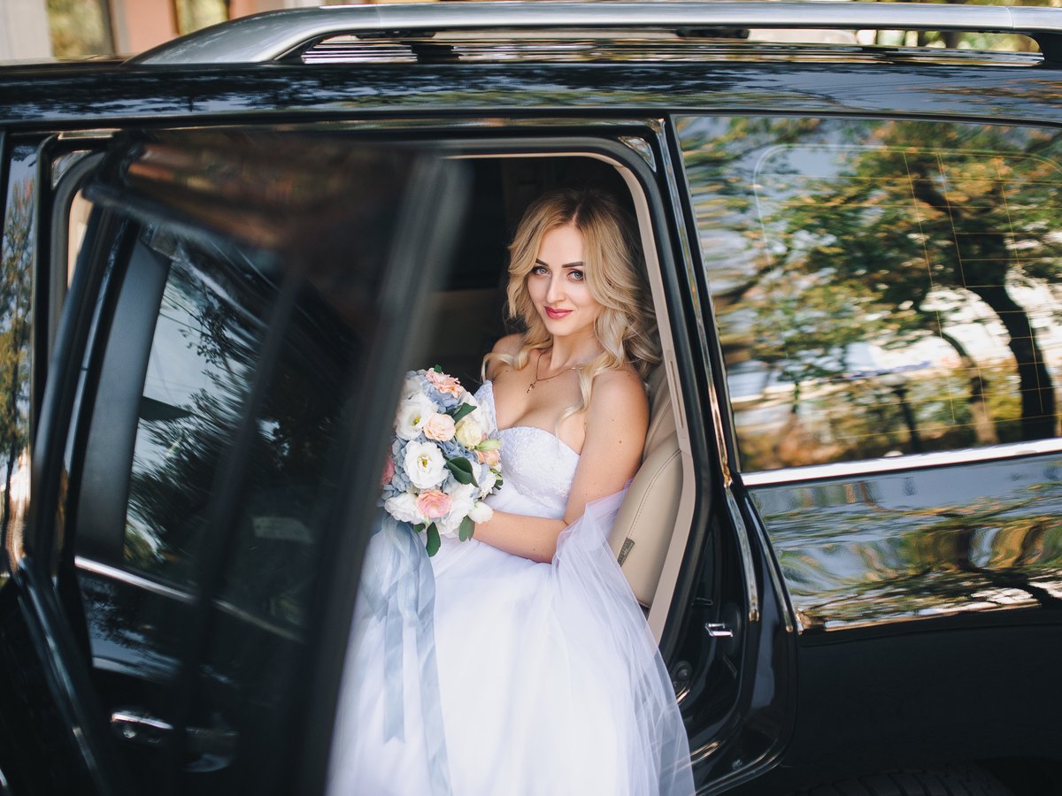 A bride standing at the open rear door of a black SUV, bouquet in hand
