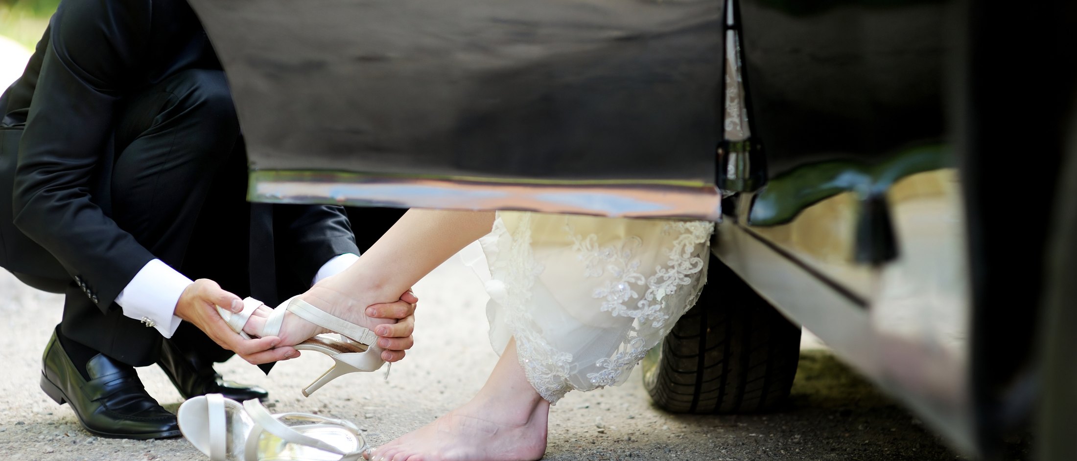 A groom kneeling to fasten a bride's shoe beside a waiting car
